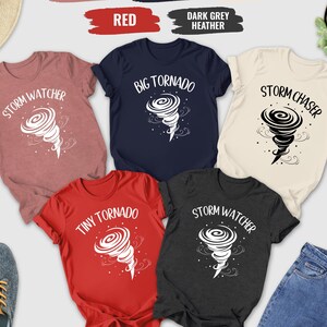 May include: A collection of t-shirts in various colors, including heather mauve, navy, red, dark grey heather, and natural. Each shirt features a white graphic of a tornado with text such as "Storm Watcher", "Big Tornado", and "Storm Chaser".