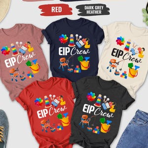 May include: A collection of short-sleeved t-shirts in various colors, including heather mauve, navy, red, dark grey heather, and natural. Each shirt features the text "EIP Crew" with colorful graphics of toys and tools. A pair of blue jeans and sneakers are also visible.