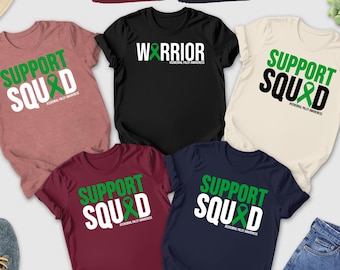 May include: A collection of t-shirts in various colors, including heather mauve, black, maroon, and navy. Each shirt features the text "SUPPORT SQUAD" with a green ribbon graphic. A black t-shirt displays "WARRIOR". Accessories include sunglasses, a hat, and jeans.
