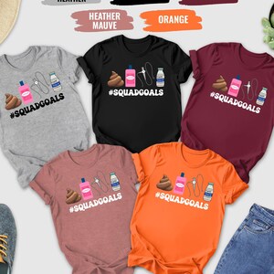 May include: A collection of t-shirts in various colors, including gray, black, maroon, orange, and heather mauve. Each shirt features a graphic design with a poop emoji, a pink bottle, a needle, and a milk carton, with the text "#SQUADGOALS".