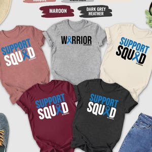 May include: A collection of t-shirts in various colors, including heather mauve, maroon, and dark grey heather. Each shirt features the words "SUPPORT SQUAD" in bold letters with a blue ribbon graphic. Other items include jeans, shoes, and a hat.