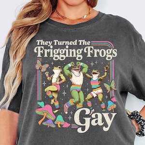 May include: Dark grey t-shirt with the text "They Turned The Frigging Frogs Gay" and a graphic of dancing frogs. The frogs are green and wearing colorful outfits. Mushrooms and butterflies are also featured in the design.
