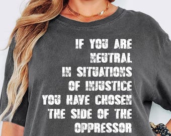 May include: A dark gray t-shirt with the quote "IF YOU ARE NEUTRAL IN SITUATIONS OF INJUSTICE YOU HAVE CHOSEN THE SIDE OF THE OPPRESSOR - DESMOND TUTU" in white text. The shirt is a relaxed fit.