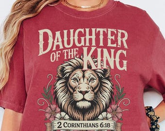 May include: Crimson-colored t-shirt with the text "DAUGHTER OF THE KING" above a lion's head illustration. Below the lion is the text "2 Corinthians 6:18" on a banner with floral accents. The shirt is a relaxed fit.
