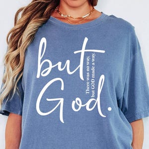 May include: Blue jean colored t-shirt with the words "but God" in white script. The text "There was no way, but GOD made a way" is printed vertically on the right side of the shirt. The shirt is a comfortable, casual style.