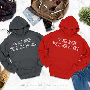 two hooded sweatshirts with humorous text, laid out on a marble surface alongside other items such as a backpack, sunglasses, and a plant.