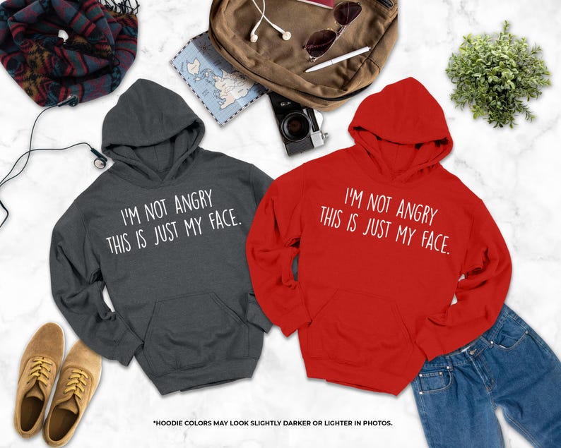 two hooded sweatshirts with humorous text, laid out on a marble surface alongside other items such as a backpack, sunglasses, and a plant.