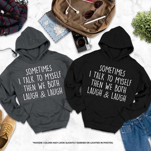 two hooded sweatshirts with humorous text printed on them, laid out on a marble surface alongside various personal items such as a backpack, a plant, and a pair of shoes.