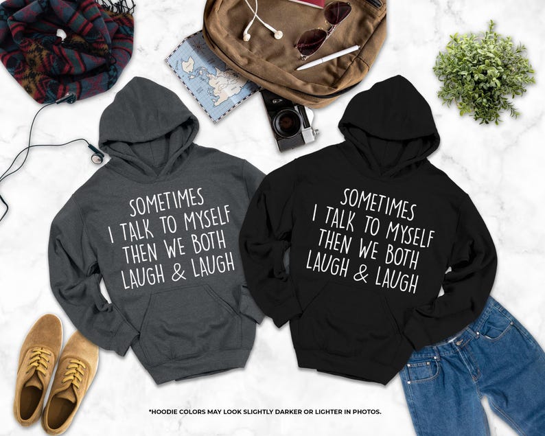 two hooded sweatshirts with humorous text printed on them, laid out on a marble surface alongside various personal items such as a backpack, a plant, and a pair of shoes.