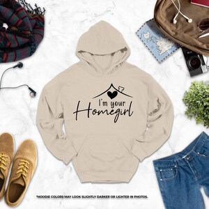 a casual outfit consisting of a beige hoodie with the text "I'm your Homegirl" printed on it, along with a pair of jeans, a pair of brown shoes, and a pair of sunglasses. The items are arranged on a marble surface, creating a visually appealing and organized display.