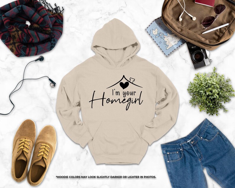 a casual outfit consisting of a beige hoodie with the text "I'm your Homegirl" printed on it, along with a pair of jeans, a pair of brown shoes, and a pair of sunglasses. The items are arranged on a marble surface, creating a visually appealing and organized display.