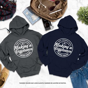 a pair of gray and navy blue hooded sweatshirts with white text, laid out on a marble surface alongside other items such as a backpack, a pair of shoes, and a plant.