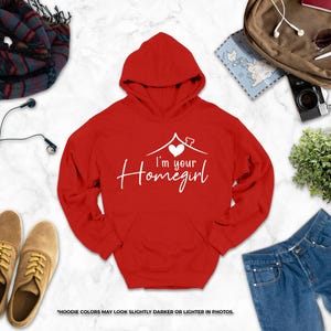 a variety of clothing and accessories, including a red hoodie with the text "I'm your Homegirl" printed on it, a pair of brown shoes, a pair of blue jeans, a scarf, a camera, and a plant.