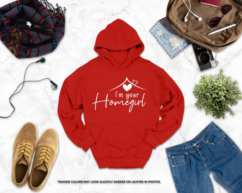 a variety of clothing and accessories, including a red hoodie with the text "I'm your Homegirl" printed on it, a pair of brown shoes, a pair of blue jeans, a scarf, a camera, and a plant.
