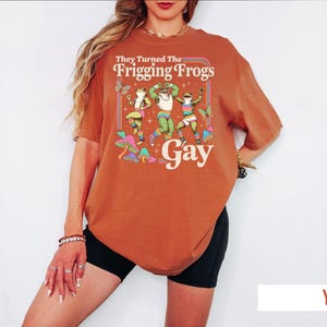 A woman wearing a red t-shirt with a graphic design of a frog character and the text "They Turned The Friggling Frogs Gay" printed on it.
