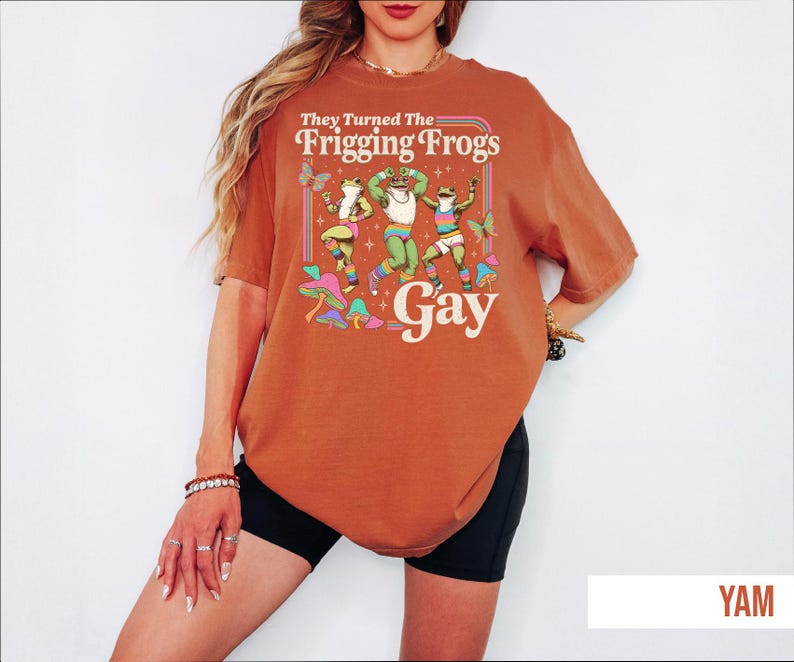 A woman wearing a red t-shirt with a graphic design of a frog character and the text "They Turned The Friggling Frogs Gay" printed on it.