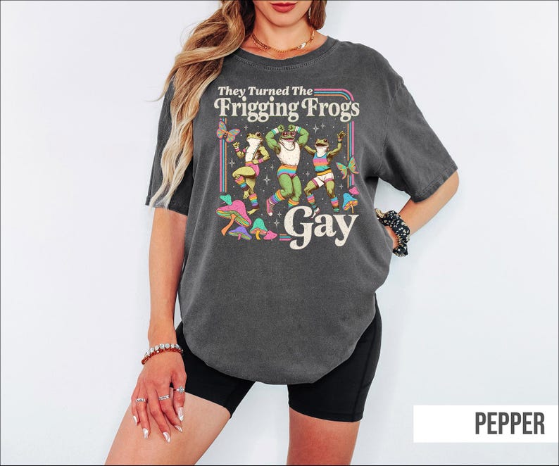 A woman wearing a gray t-shirt with a colorful graphic design featuring a frog and the text "They Turned The Friggling Frogs Gay".