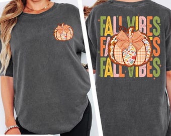 May include: A dark gray t-shirt with a pumpkin graphic on the front and back. The front features a small pumpkin with a bow. The back of the shirt has the words "FALL VIBES" and a larger pumpkin design. The pumpkin designs have a plaid and floral pattern.