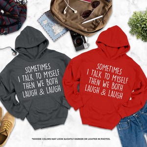 two hooded sweatshirts with humorous text printed on them, laid out on a marble surface alongside various personal items such as a camera, headphones, and a plant.