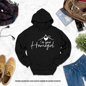 a variety of clothing and accessories, including a black hoodie with the text "I'm your Homegirl" printed on it, a pair of brown shoes, a pair of blue jeans, a scarf, a camera, and a plant.