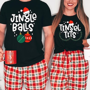 May include: Matching black Christmas t-shirts with humorous text and festive graphics. One shirt says "Jingle Balls" with ornaments, the other says "Tinsel Tits" with a wreath. Both wear red plaid pajama pants and hold mugs.