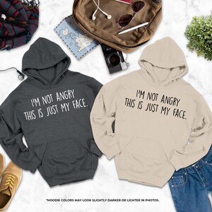 two hooded sweatshirts with humorous text printed on them, laid out on a marble surface alongside other items such as a camera, sunglasses, and a plant.