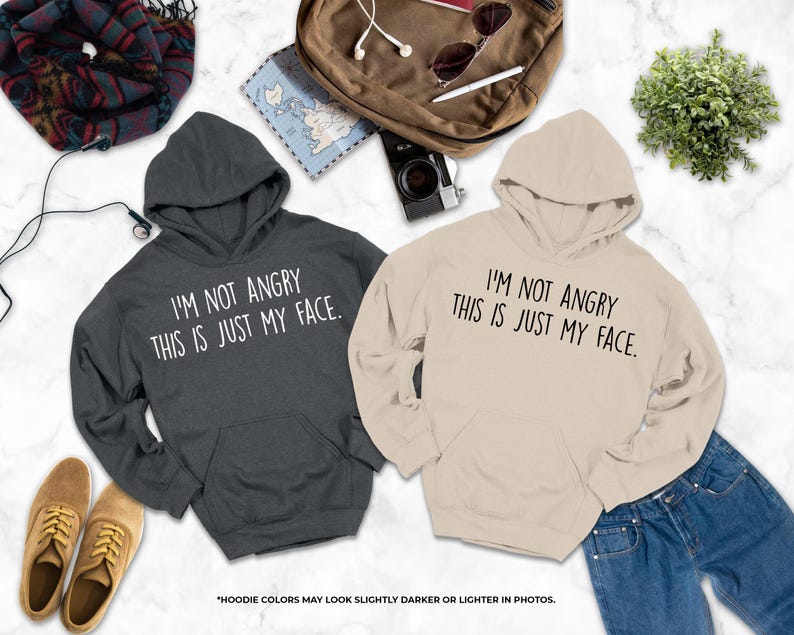 two hooded sweatshirts with humorous text printed on them, laid out on a marble surface alongside other items such as a camera, sunglasses, and a plant.