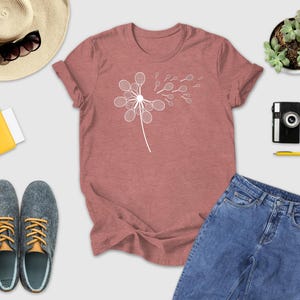 a casual outfit consisting of a pink t-shirt with a white flower design, a pair of blue jeans, gray shoes, a straw hat, a yellow wallet, a camera, a pen, and a potted plant.