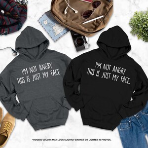 two hooded sweatshirts with humorous text, laid out on a marble surface alongside various personal items such as a backpack, a plant, and a pair of shoes.