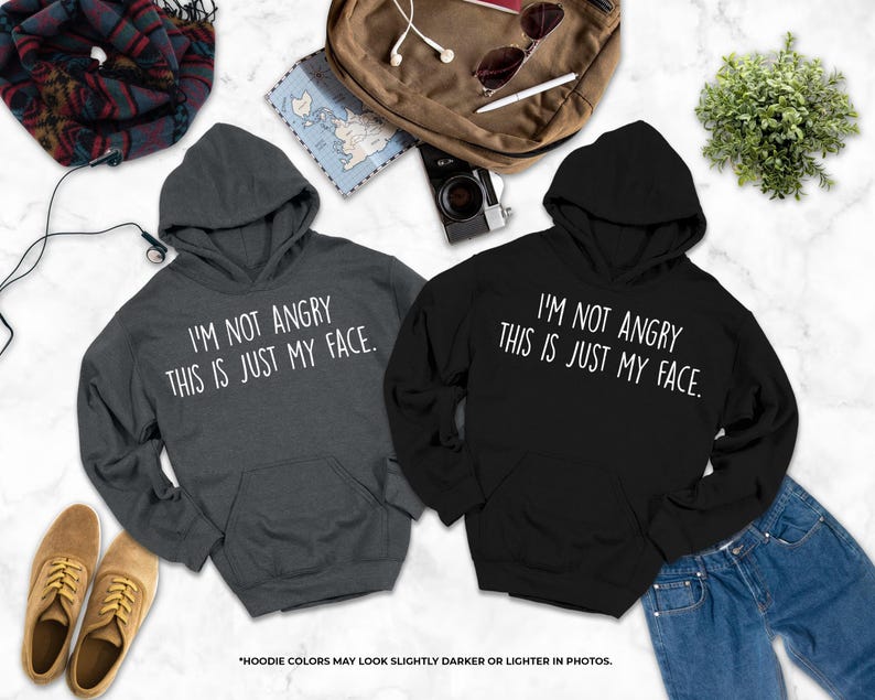 two hooded sweatshirts with humorous text, laid out on a marble surface alongside various personal items such as a backpack, a plant, and a pair of shoes.