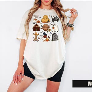 A woman wearing a white t-shirt with a graphic design of various Halloween-themed items, including a skeleton, a pumpkin, a ghost, and a barn.