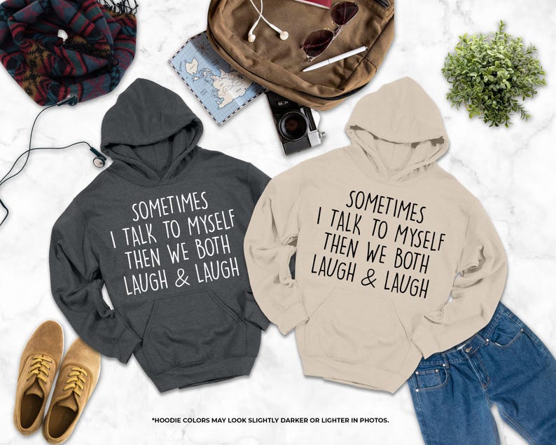 two hooded sweatshirts with humorous text printed on them, laid out on a marble surface alongside various personal items such as a camera, headphones, and a plant.