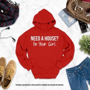 a red hoodie with the text "NEED A HOUSE? I'M YOUR GIRL" printed on it, along with various clothing items such as jeans, a pair of shoes, and a scarf. There are also accessories like earphones, a camera, and sunglasses. The image also includes a map and a plant, adding to the overall composition.