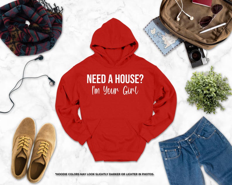 a red hoodie with the text "NEED A HOUSE? I'M YOUR GIRL" printed on it, along with various clothing items such as jeans, a pair of shoes, and a scarf. There are also accessories like earphones, a camera, and sunglasses. The image also includes a map and a plant, adding to the overall composition.