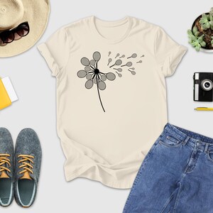a casual outfit consisting of a white t-shirt with a graphic design, a pair of blue jeans, gray shoes, a straw hat, sunglasses, a yellow wallet, a camera, a pen, and a potted plant.