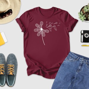 a variety of casual clothing and accessories, including a burgundy t-shirt with a white flower design, a pair of jeans, a pair of sneakers, a hat, sunglasses, a camera, a yellow notebook, a yellow pen, a black camera, a black wallet, a yellow folder, and a green plant.