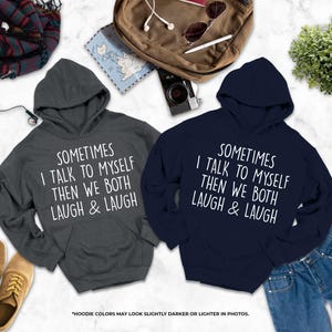 two hooded sweatshirts with humorous text printed on them, laid out on a marble surface alongside various personal items such as a backpack, a pair of shoes, and a plant.