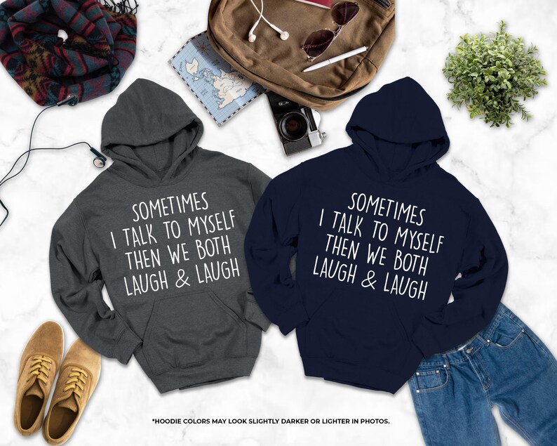 two hooded sweatshirts with humorous text printed on them, laid out on a marble surface alongside various personal items such as a backpack, a pair of shoes, and a plant.