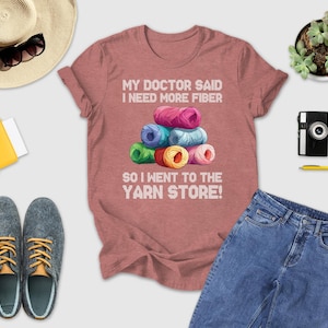 a pink t-shirt with a humorous text, a stack of colorful yarn, a hat, sunglasses, a camera, a wallet, a pair of shoes, a pen, a book, and a plant.