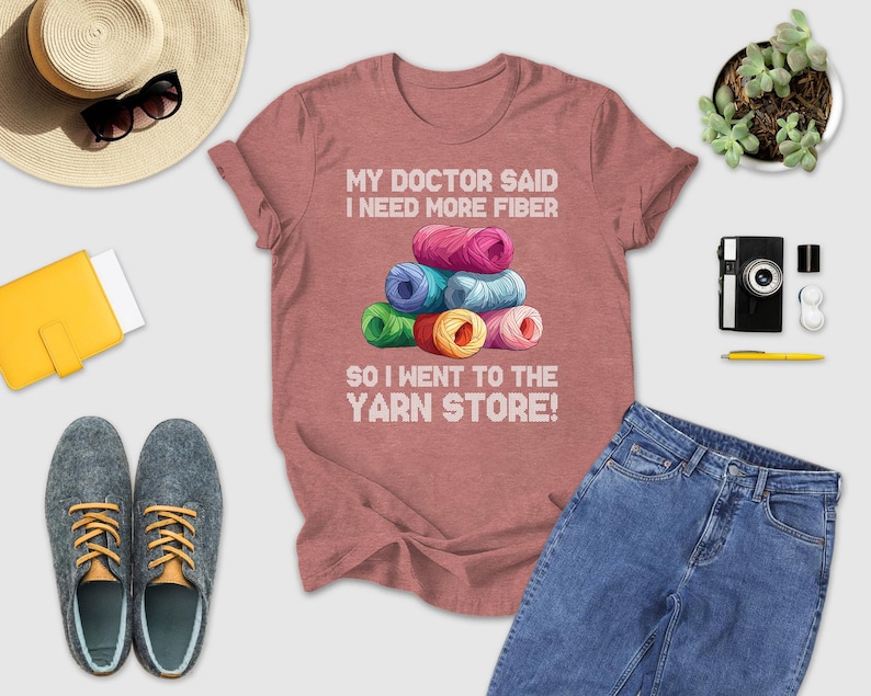 a pink t-shirt with a humorous text, a stack of colorful yarn, a hat, sunglasses, a camera, a wallet, a pair of shoes, a pen, a book, and a plant.