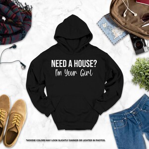 a black hoodie with the text "NEED A HOUSE? I'M YOUR GIRL" printed on it, along with various clothing items such as jeans, a pair of shoes, and a backpack.