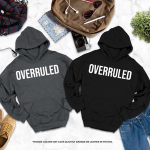 two gray hooded sweatshirts with the word "OVERRULED" printed on them, along with a pair of jeans, a pair of shoes, and some other items such as a backpack, a map, and a plant.