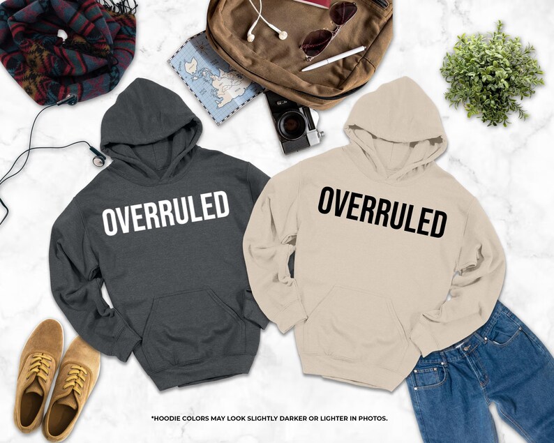 two hooded sweatshirts with the word "OVERRULED" printed on them, along with a pair of jeans, a pair of shoes, and some other items such as a backpack, a camera, and a plant.