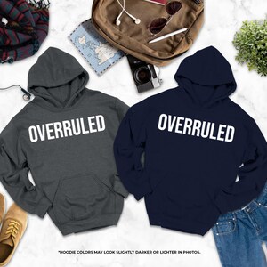 two gray hooded sweatshirts with the word "OVERRULED" printed on them, along with a pair of jeans, a pair of shoes, and some other items such as a backpack, a map, and a plant.