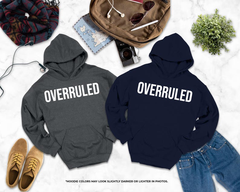 two gray hooded sweatshirts with the word "OVERRULED" printed on them, along with a pair of jeans, a pair of shoes, and some other items such as a backpack, a map, and a plant.