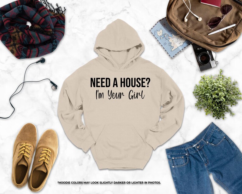 a casual outfit consisting of a beige hoodie with the text "NEED A HOUSE? I'M YOUR GIRL" printed on it, along with a pair of jeans, a pair of brown shoes, a scarf, a camera, and a plant.