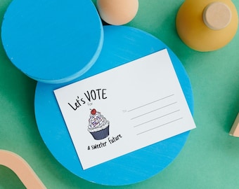 Puede incluir: Una postal blanca con el texto "Let's Vote for a sweeter future" y una ilustración de cupcake. La postal está sobre una plataforma circular azul, con una esfera marrón claro y un objeto amarillo con una parte superior de madera. El fondo es verde azulado.