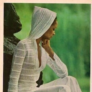 Puede incluir: Una blusa blanca de ganchillo con capucha y un profundo escote en V. La blusa se lleva con pantalones blancos y la modelo está sentada en una roca.