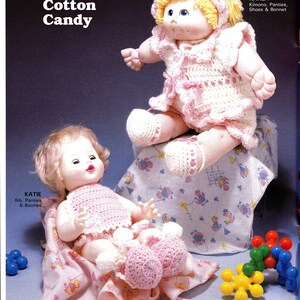 May include: Two crocheted dolls dressed in pink and white outfits. The doll on the left is named Katie and is wearing a bib, panties, and booties. The doll on the right is named Karla and is wearing a kimono, panties, shoes, and a bonnet. The dolls are sitting on a pink and white blanket with a colorful toy on the right.