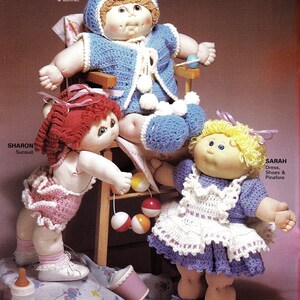 May include: Three Cabbage Patch Kids dolls wearing crocheted outfits. The doll in the middle is sitting in a wooden chair. The doll on the left is wearing a pink sunsuit and the doll on the right is wearing a purple dress with a white crocheted pinafore. The dolls are playing with a colorful toy.
